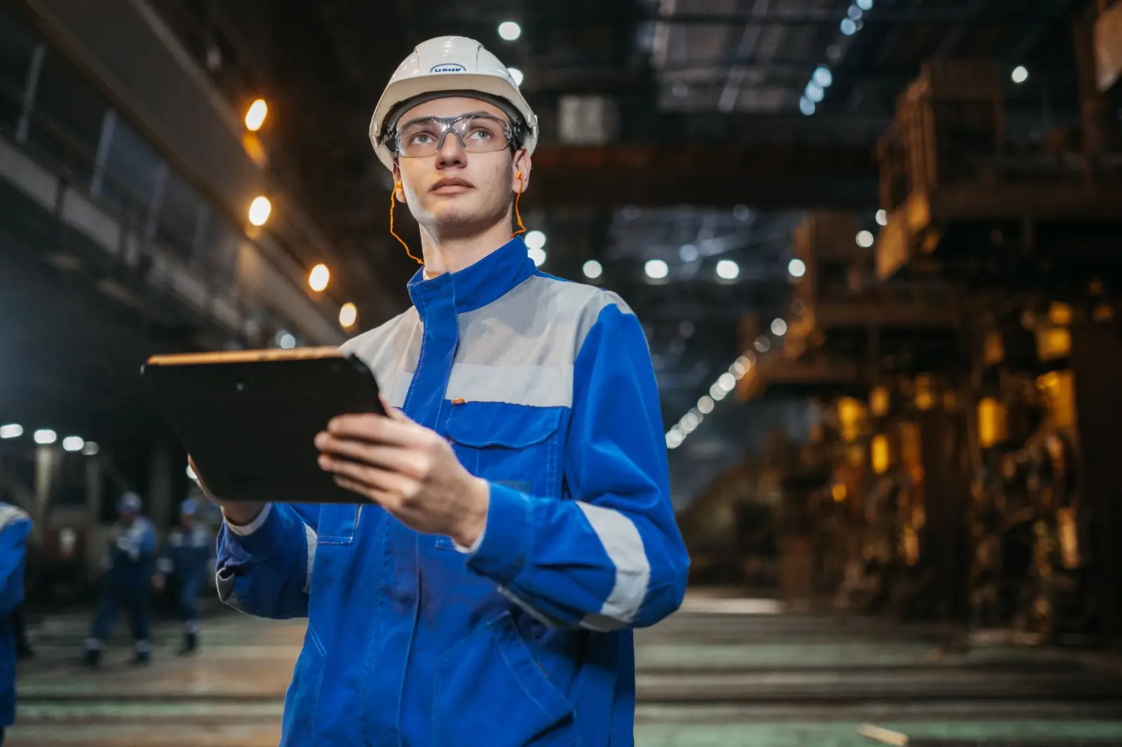Mitarbeiter in Arbeitskleidung prüft Daten auf einem Tablet in einer großen Produktionshalle